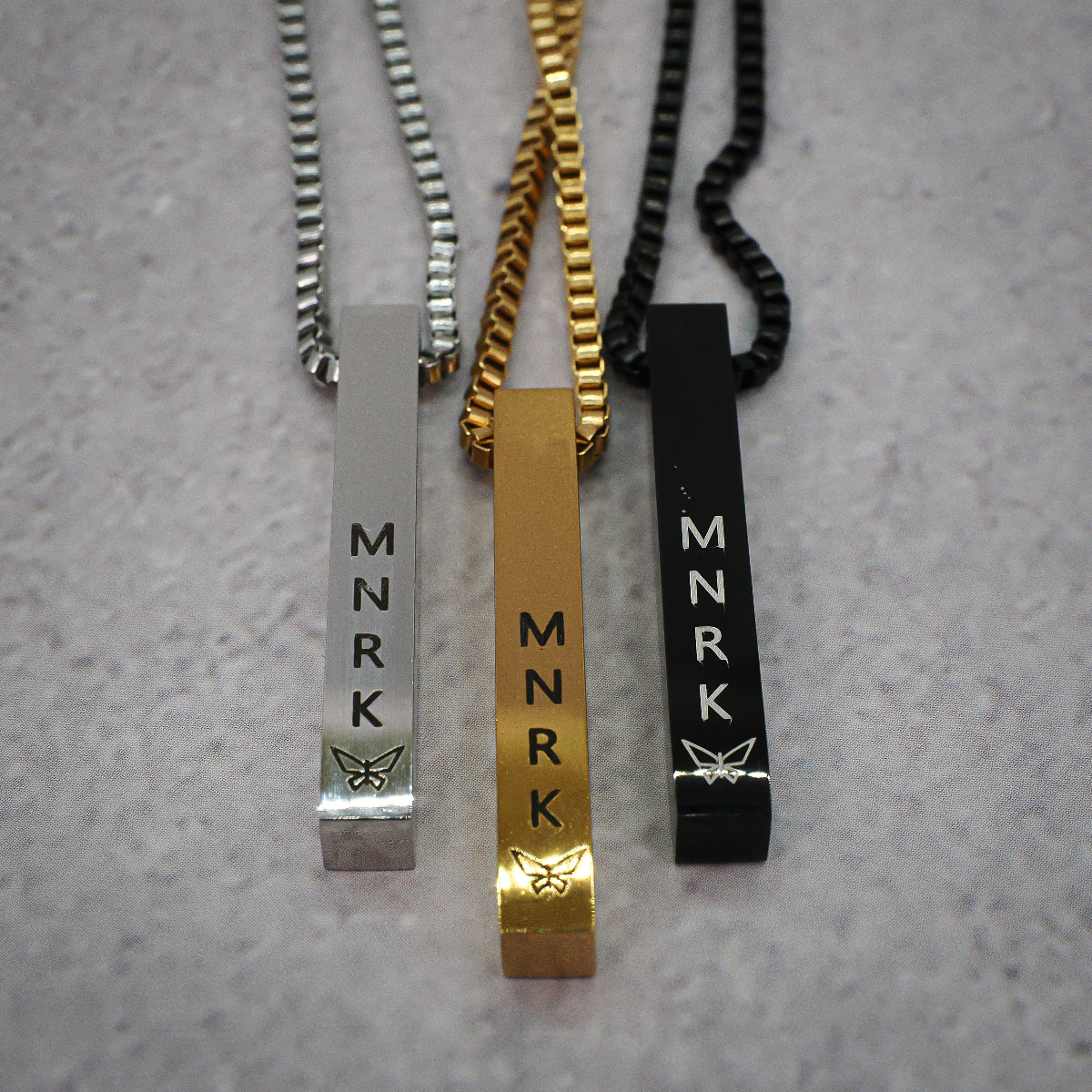 Three necklaces (silver, black and gold) with bar pendants displaying 'MNRK' branding on a grey counter.