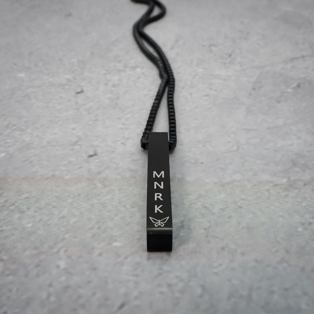 Black necklace with bar pendant displaying 'MNRK' branding on a grey counter.