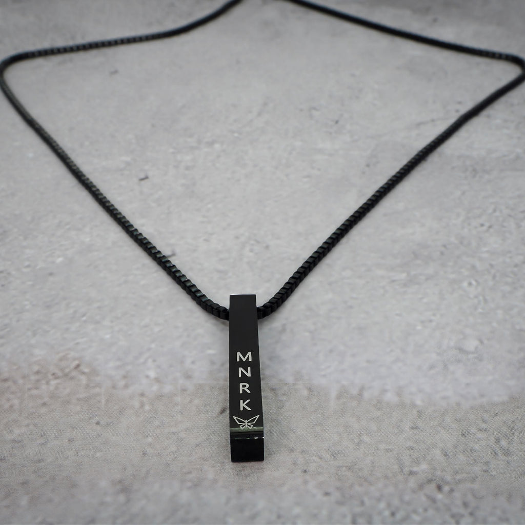 Black necklace with bar pendant displaying 'MNRK' branding on a grey counter. The chain lays around it.