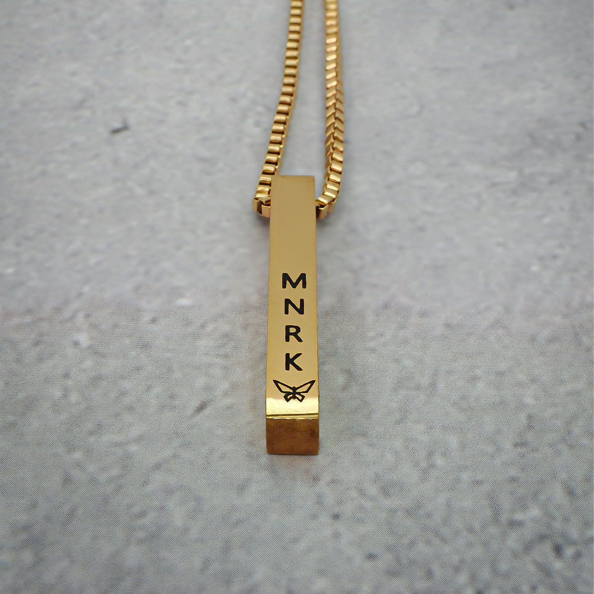 Gold necklace with bar pendant displaying 'MNRK' branding on a grey counter.