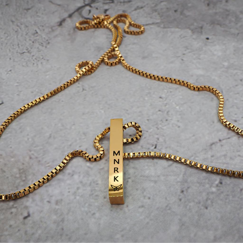 Gold necklace with bar pendant displaying 'MNRK' branding on a grey counter. The chain lays around it