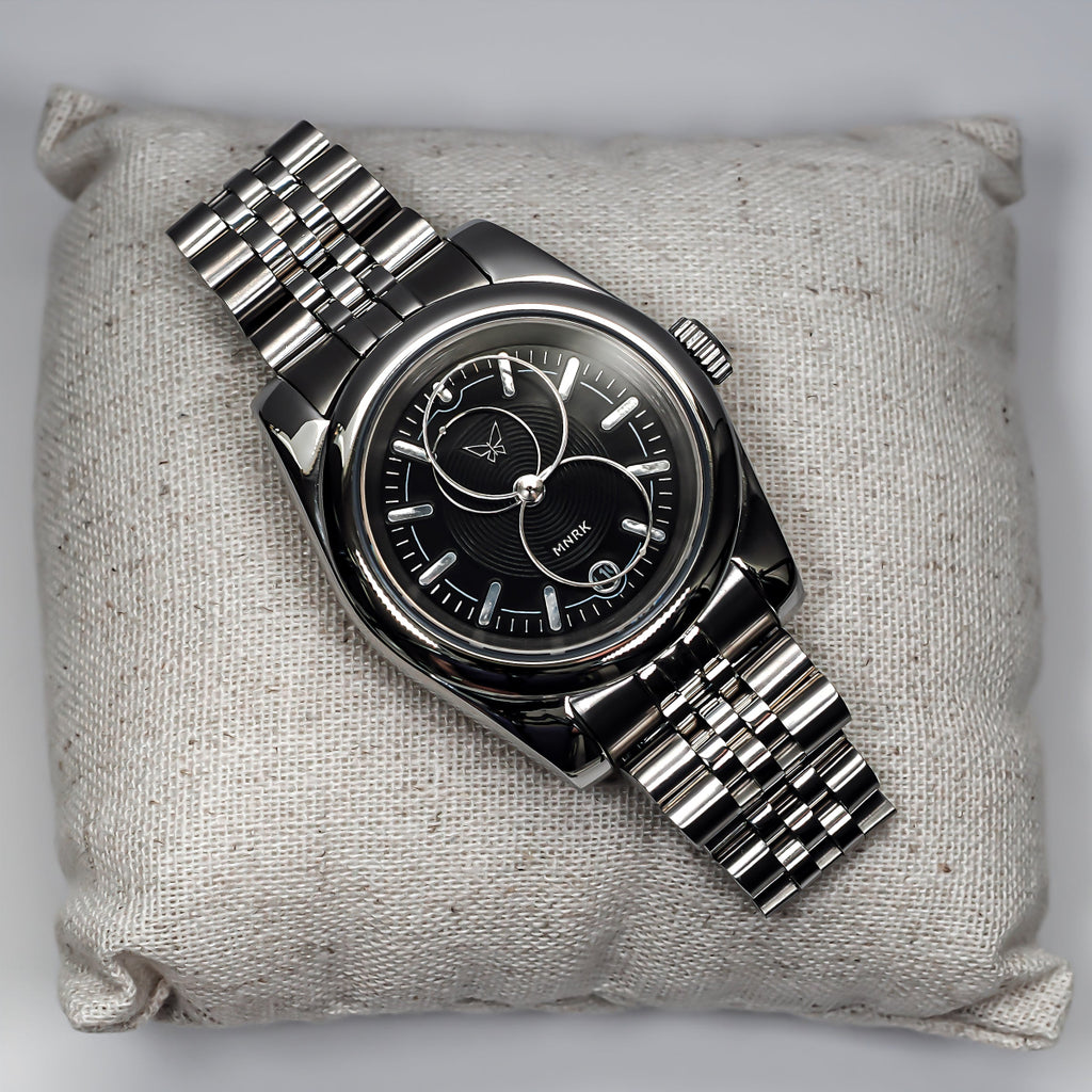 Luxury silver watch, with a black dial and circular hands, viewed from the front. It lays upon a small pillow