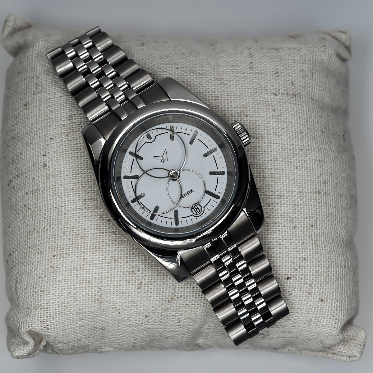 Luxury silver watch, with a white dial and circular hands, viewed from the front. It lays upon a small pillow
