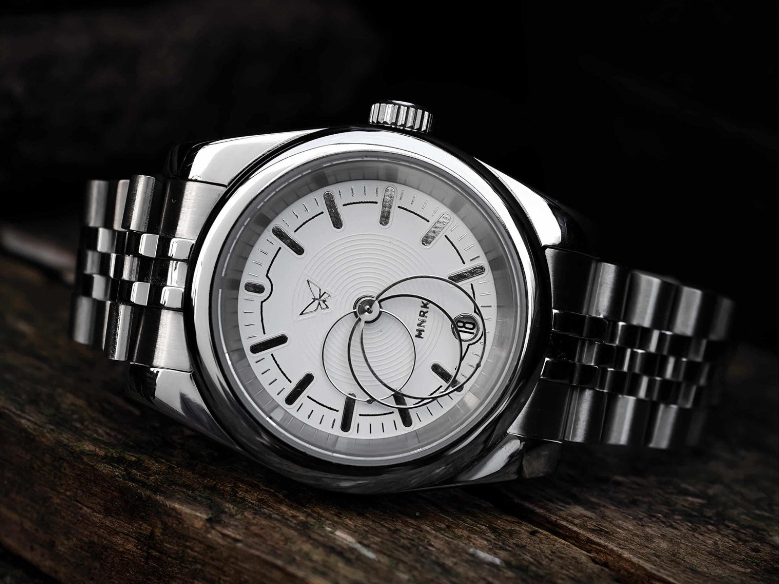 Luxury silver watch with a white dial and circular hands on a dark background, it rests on its side 