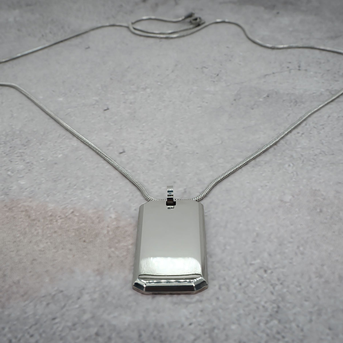 Silver rectangular pendant on a chain on a grey counter.