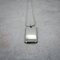 Silver rectangular pendant on a chain on a grey counter, with the chain straightened.