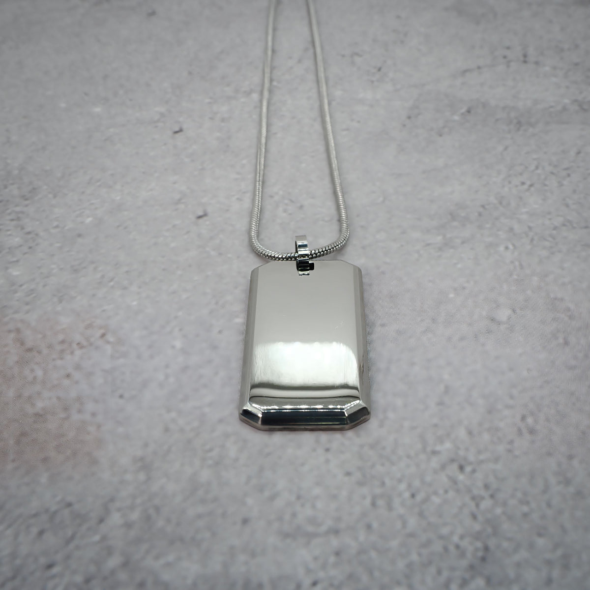 Silver rectangular pendant on a chain on a grey counter, with the chain straightened.