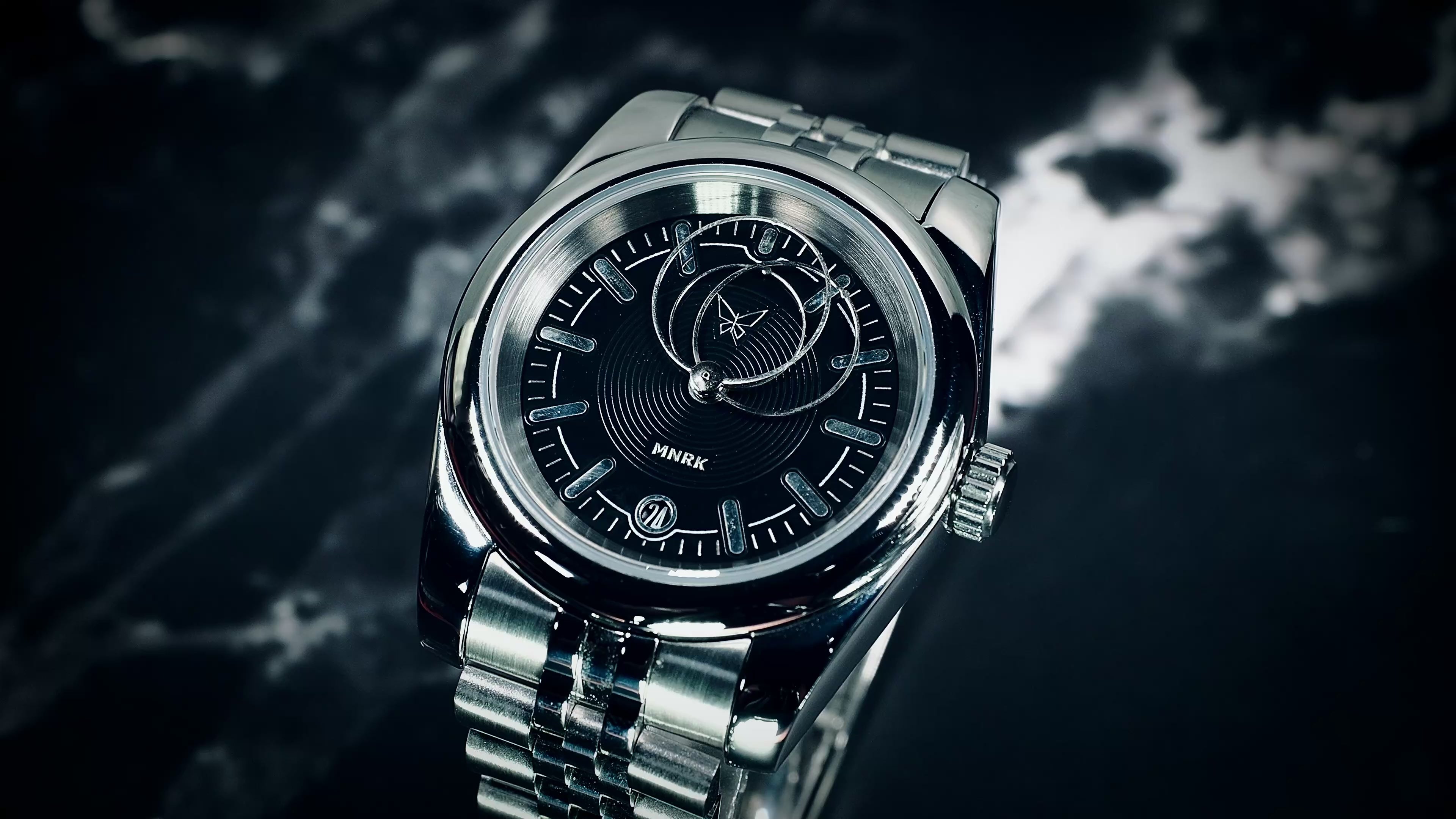 Luxury silver watch, with a black dial and circular hands, viewed from the front. The video shows the hands in motion
